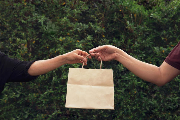Woman taking an empty brown recycled paper shopping bag from a man with green leaves in the background.